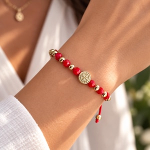 May include: A beaded bracelet featuring alternating bright red faceted beads and smooth gold-toned metallic spheres. A central circular gold-toned charm with an embossed intricate pattern serves as the focal point. The bracelet includes a red cord.