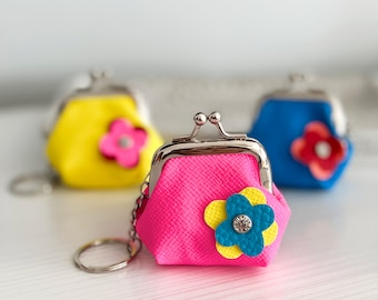 Puede incluir: Tres pequeñas carteras de monedas con detalles de flores. Las carteras son amarillas, rosas y azules. Cada cartera tiene un cierre plateado y un llavero.