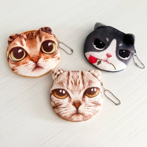 May include: Three plush cat coin purses with zipper closures and metal keyrings. The purses feature different cat faces with large eyes and are brown, black, and tan.