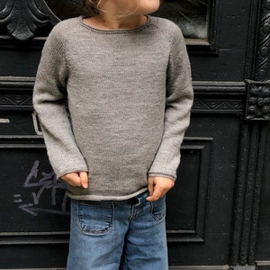 Könnte beinhalten: Ein Kind trägt einen grauen Strickpullover und eine blaue Jeans mit weitem Bein. Das Kind steht vor einer schwarzen Tür.