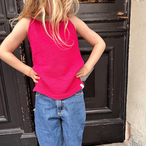 May include: A young person wearing a bright pink sleeveless knitted top and blue denim jeans. The person is standing in front of a black wooden door.