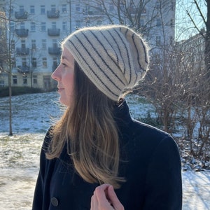 Könnte beinhalten: Eine Frau trägt einen dunkelblauen Mantel mit einer weiß-schwarz gestreiften Mütze. Sie steht in einem verschneiten Park mit einem Gebäude im Hintergrund.