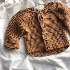 Könnte beinhalten: Ein braunes Strickjäckchen mit Holzknöpfen. Das Jäckchen hat einen runden Ausschnitt und lange Ärmel. Das Etikett auf dem Jäckchen lautet "my knit people berlin".