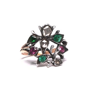 May include: A silver-toned ring with a floral design, featuring clear, green, and red gemstones. The ring has a gold-colored band and is set against a white background. The gemstones are cut in various shapes, adding to the ring's intricate appearance.