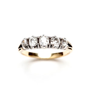 May include: A gold ring with five clear, round gemstones set in a row. The ring has a classic design with a gold band and a white background. The gemstones are evenly spaced and catch the light.