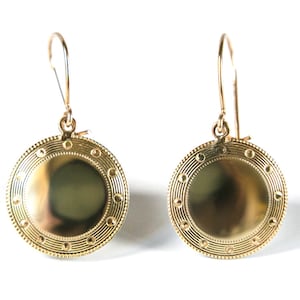 May include: A pair of gold-toned earrings with a round, disc-shaped design. The discs have a textured, circular pattern around the edge.