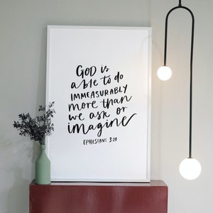 Può includere: Stampa incorniciata bianca con testo in calligrafia nera che recita "Dio è in grado di fare molto più di quanto possiamo chiedere o immaginare" con il riferimento biblico "Efesini 3:20" sotto il testo. La stampa è appesa a un muro sopra uno scaffale color borgogna con un'apertura ad arco.