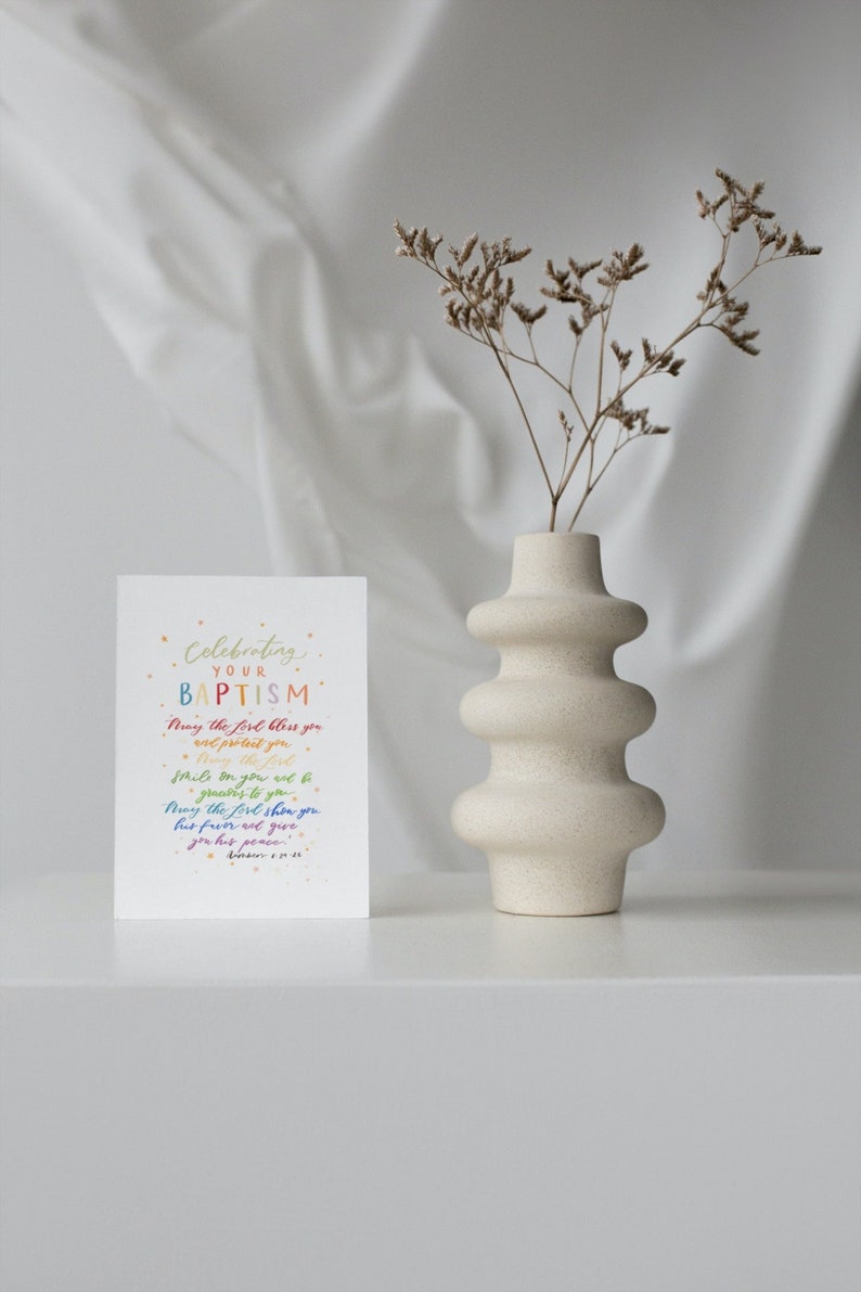 May include: A white card with a rainbow-colored message celebrating a baptism. The message reads "Celebrating your Baptism. May the Lord bless you and protect you. Smile on you and be gracious to you. May the Lord show you his favor and give you his peace." The card is sitting on a white surface next to a white vase with dried flowers.