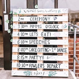 May include: A white wooden pallet sign with black handwritten text listing the schedule for a wedding. The sign reads "Naomi & Sam's wedding" at the top and includes times for the ceremony, drinks, dinner, speeches, cake cutting, dancing, and the end of the reception.
