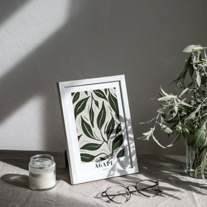 May include: A white picture frame with a green and white botanical print. The print features a leafy design with the word "AGAPE" at the bottom. The frame is sitting on a white linen surface with a glass candle jar and a pair of eyeglasses.