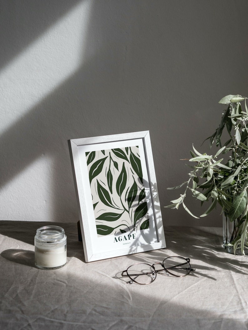 May include: A white picture frame with a green and white botanical print. The print features a leafy design with the word "AGAPE" at the bottom. The frame is sitting on a white linen surface with a glass candle jar and a pair of eyeglasses.