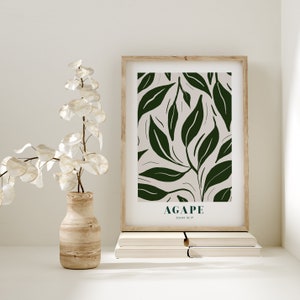May include: A framed print with a green leafy pattern and the word "AGAPE" with the scripture reference "JOHN 10:17" underneath. The print is sitting on a stack of books on a white surface.