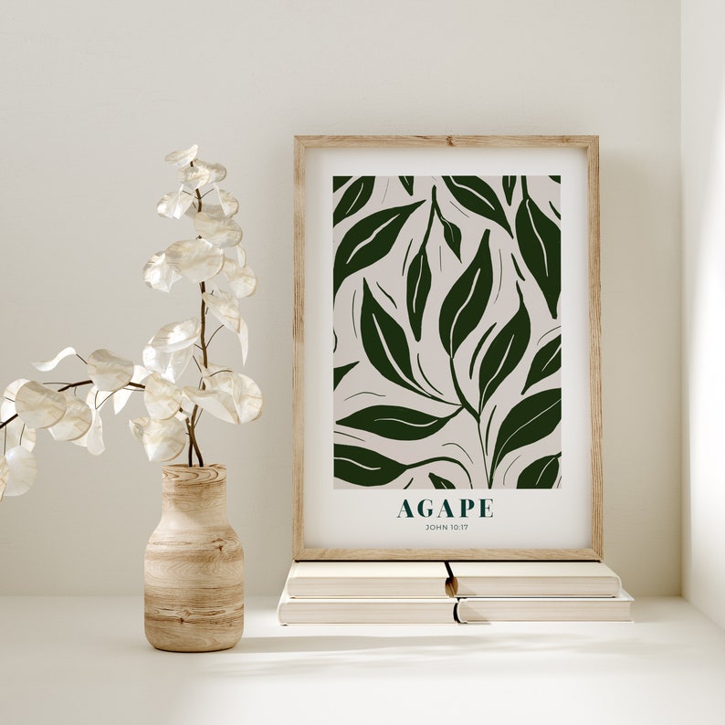 May include: A framed print with a green leafy pattern and the word "AGAPE" with the scripture reference "JOHN 10:17" underneath. The print is sitting on a stack of books on a white surface.
