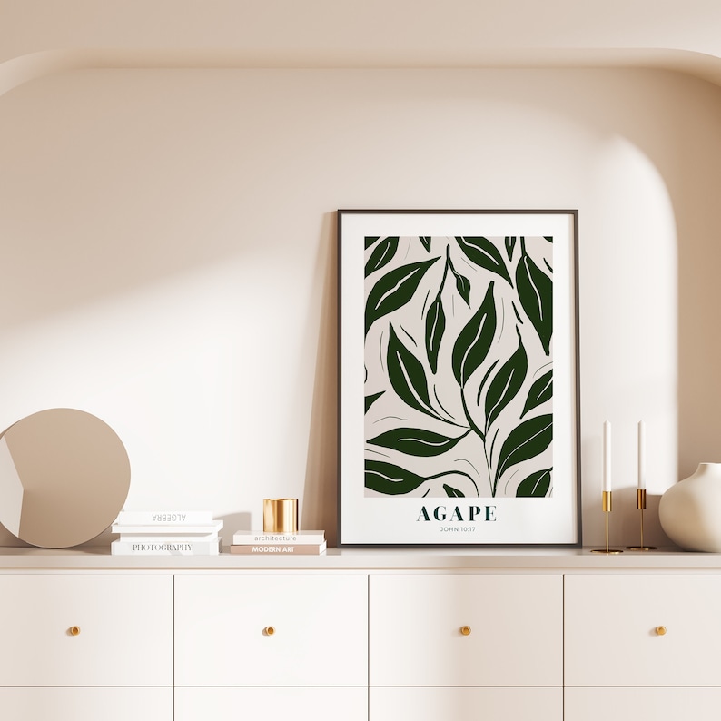 May include: A framed print with a dark green leafy pattern on a cream background. The print has the word "AGAPE" in black text and the text "JOHN 13:17" in smaller black text below it. The print is hanging on a white wall above a white dresser with three drawers. There are three books stacked on the dresser, a gold candle holder, and two white candles.
