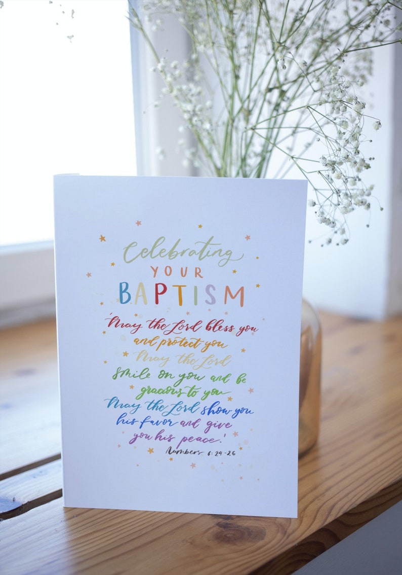 May include: A white card with a colorful handwritten message celebrating a baptism. The message reads "Celebrating your Baptism. May the Lord bless you and protect you. May the Lord smile on you and be gracious to you. May the Lord show you his favor and give you his peace! Numbers 6:29-26."