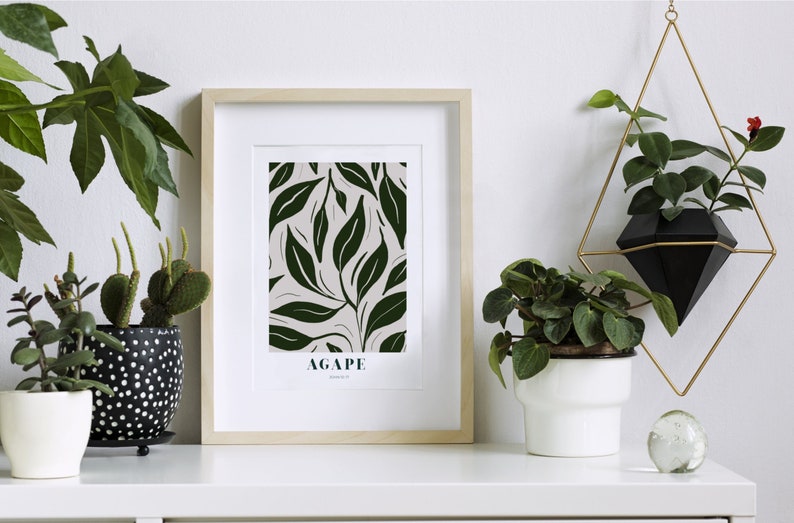 May include: A framed print with a green leaf pattern and the word "AGAPE" in black text. The print is on a white shelf with several potted plants, including a cactus and a hanging plant in a black diamond-shaped planter.