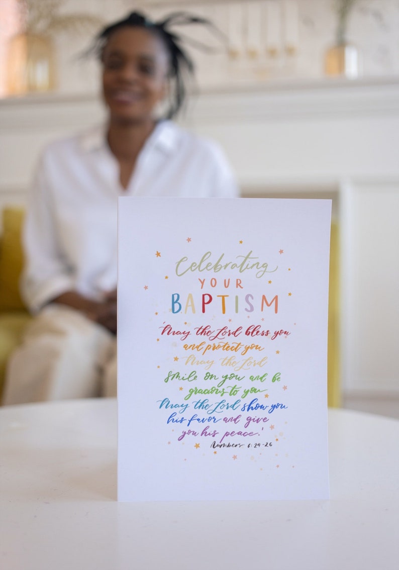 May include: A white card with a rainbow-colored message celebrating a baptism. The message reads "Celebrating Your Baptism. May the Lord bless you and protect you. May the Lord smile on you and be gracious to you. May the Lord show you his favor and give you his peace! Numbers 6:24-26."
