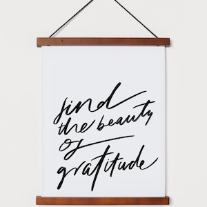 Può includere: Una stampa su tela bianca con calligrafia nera che recita "find the beauty of gratitude" appesa a una parete bianca con una cornice in legno.