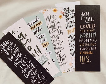 May include: A collection of inspirational bookmarks with handwritten calligraphy. The bookmarks feature various quotes and phrases in white against backgrounds of black, light blue, and floral patterns. The text includes phrases like "Your word is a lamp unto my feet" and "You are loved."