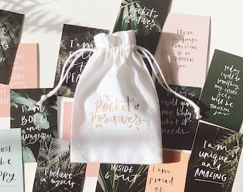 Puede incluir: Una bolsa de tela blanca con cordón de ajuste, con el texto "Pocket Positives" impreso en letras doradas. La bolsa está llena de pequeñas tarjetas con afirmaciones positivas impresas en ellas. Las tarjetas tienen una variedad de fondos, incluyendo hojas verdes y rosa.