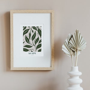 May include: A framed print with a green leaf pattern and the word "AGAPE" in white text. The print is hanging on a light brown wall next to a white vase with dried palm leaves.