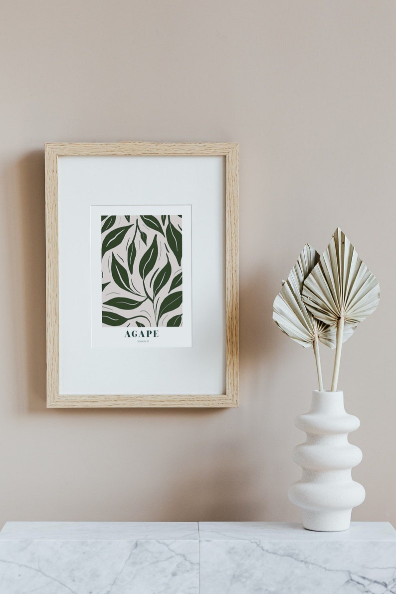May include: A framed print with a green leaf pattern and the word "AGAPE" in white text. The print is hanging on a light brown wall next to a white vase with dried palm leaves.
