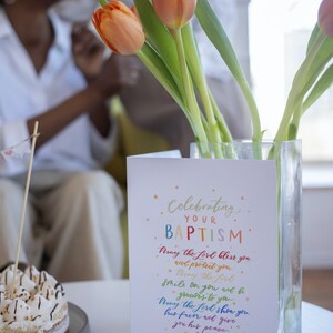 May include: A white card with colorful text that reads "Celebrating Your Baptism." The card has a rainbow-colored message that reads "May the Lord bless you and protect you. May the Lord smile on you and be gracious to you. May the Lord show you his favor and give you his peace! Numbers 6:24-26." The card is sitting on a table with a cake and a vase of orange tulips.