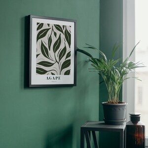 May include: A black framed print with a white background and a green leaf pattern. The print has the word "AGAPE" in black text at the bottom. The print is hanging on a green wall next to a potted plant and a small black side table.