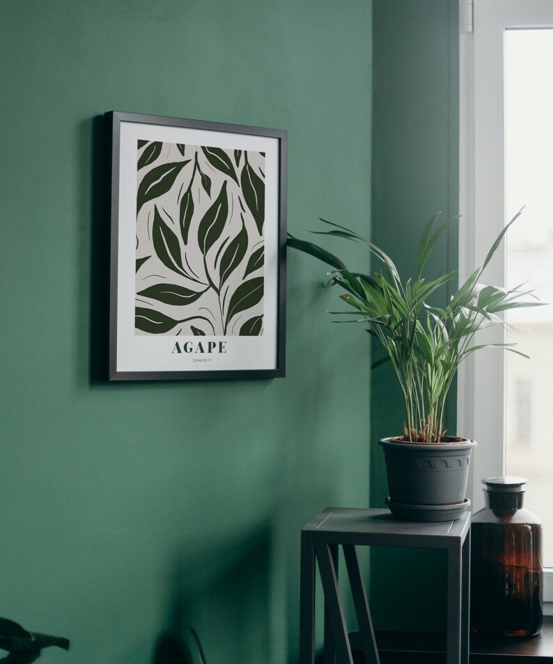 May include: A black framed print with a white background and a green leaf pattern. The print has the word "AGAPE" in black text at the bottom. The print is hanging on a green wall next to a potted plant and a small black side table.