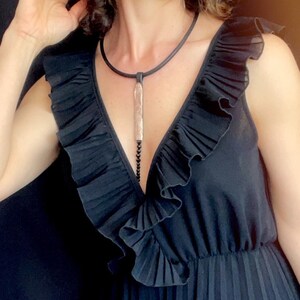 May include: A woman wearing a black sleeveless dress with a ruffled neckline and a long pendant necklace. The dress has a pleated skirt and is made of a soft, flowing fabric.