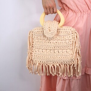 May include: A crocheted cream-colored tote bag with a light yellow wooden handle and fringe detailing.