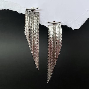 Puede incluir: Un par de pendientes de cadena de plata con un diseño de flecos. Los pendientes cuelgan de un trozo de papel blanco sobre un fondo negro.