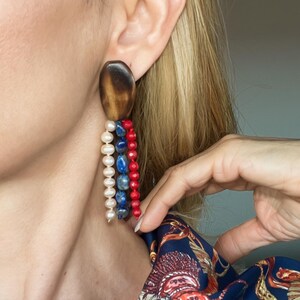 Peut inclure: Une boucle d'oreille composée d'un grand clou ovale en bois brun foncé lisse. Trois brins pendants sont fixés au clou, comprenant une rangée de perles blanches, une rangée de perles en pierre bleue et une rangée de petites perles rouges vives.