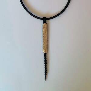 May include: A black choker necklace with gold accents and a pendant made of a gold-colored bar with a string of black beads hanging from it.