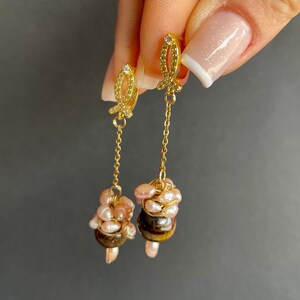 May include: Gold dangle earrings with a small fish charm and a cluster of pink pearls and a tiger's eye stone at the bottom.