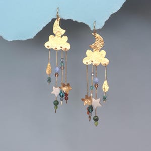 Peut inclure: Boucles d'oreilles dorées avec un motif de nuage et de croissant de lune. Chaque boucle d'oreille est ornée d'un nuage et d'une lune, avec des perles pendantes et des étoiles. Elles sont conçues pour être portées en paire.