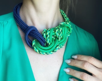 May include: A green and blue rope necklace with a knotted design. The necklace is worn by a person wearing a green top.