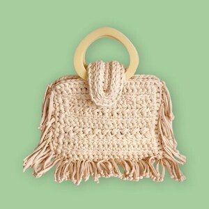 May include: A beige crocheted bag with a wooden handle and fringe detailing. The bag is on a light green background.