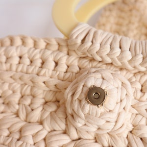 May include: A close-up of a cream-colored crocheted bag with a large, round, cream-colored handle and a gold-colored button.