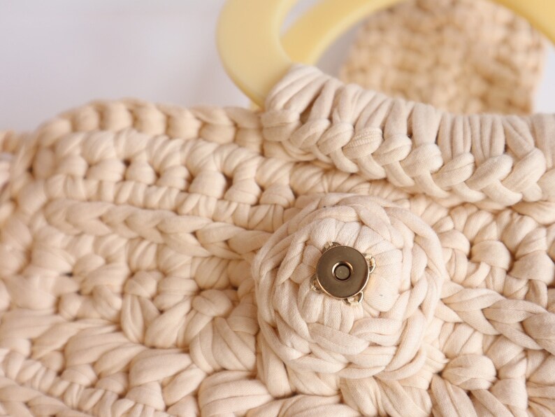 May include: A close-up of a cream-colored crocheted bag with a large, round, cream-colored handle and a gold-colored button.