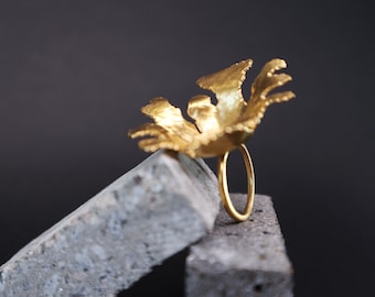 May include: A gold ring with a large, textured, flower-shaped design. The ring is sitting on two gray concrete blocks.