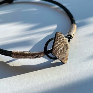 May include: A black leather choker necklace with a gold-toned geometric pendant. The pendant has a textured surface and is shaped like a diamond.