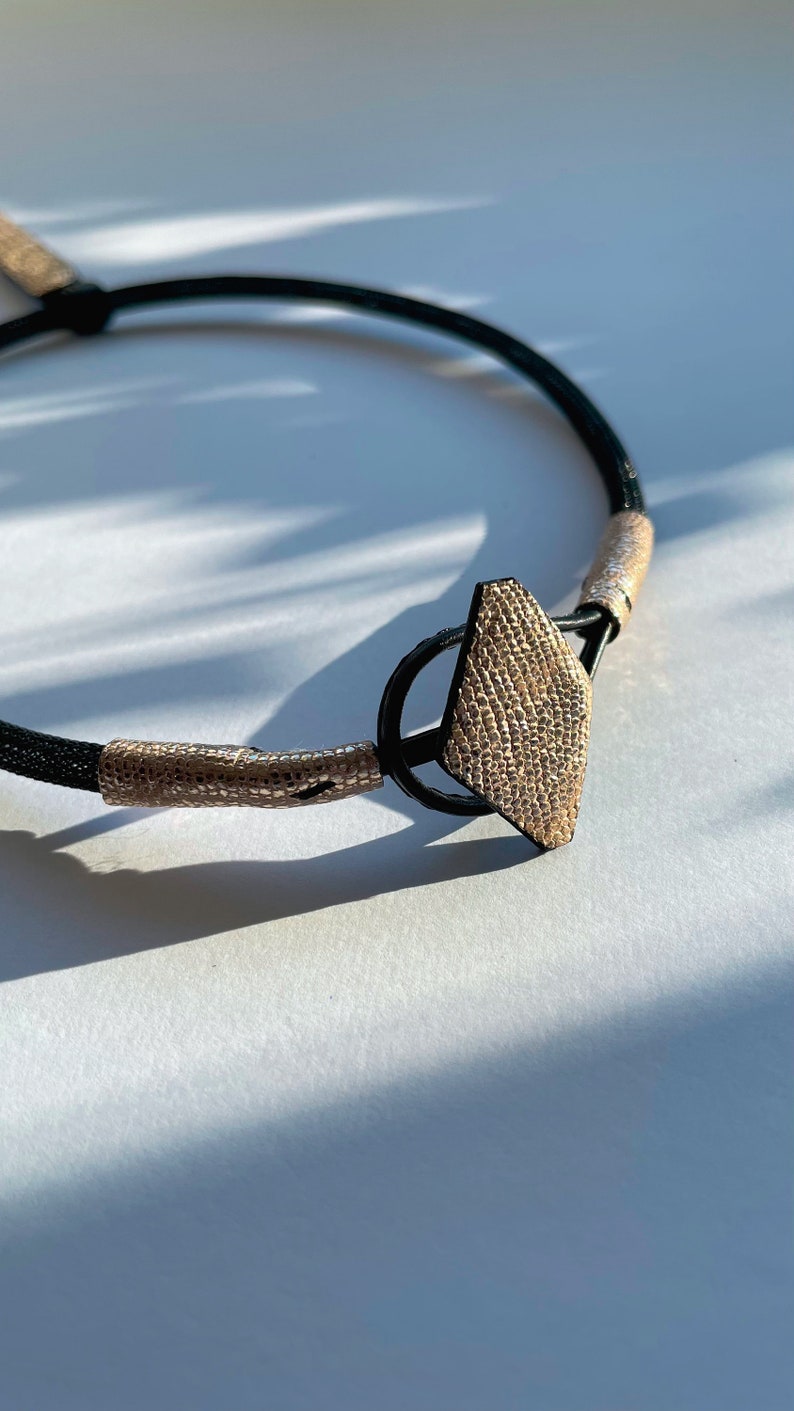 May include: A black leather choker necklace with a gold-toned geometric pendant. The pendant has a textured surface and is shaped like a diamond.