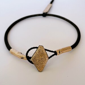 May include: A black cord bracelet with a gold diamond-shaped clasp. The clasp has a textured surface.