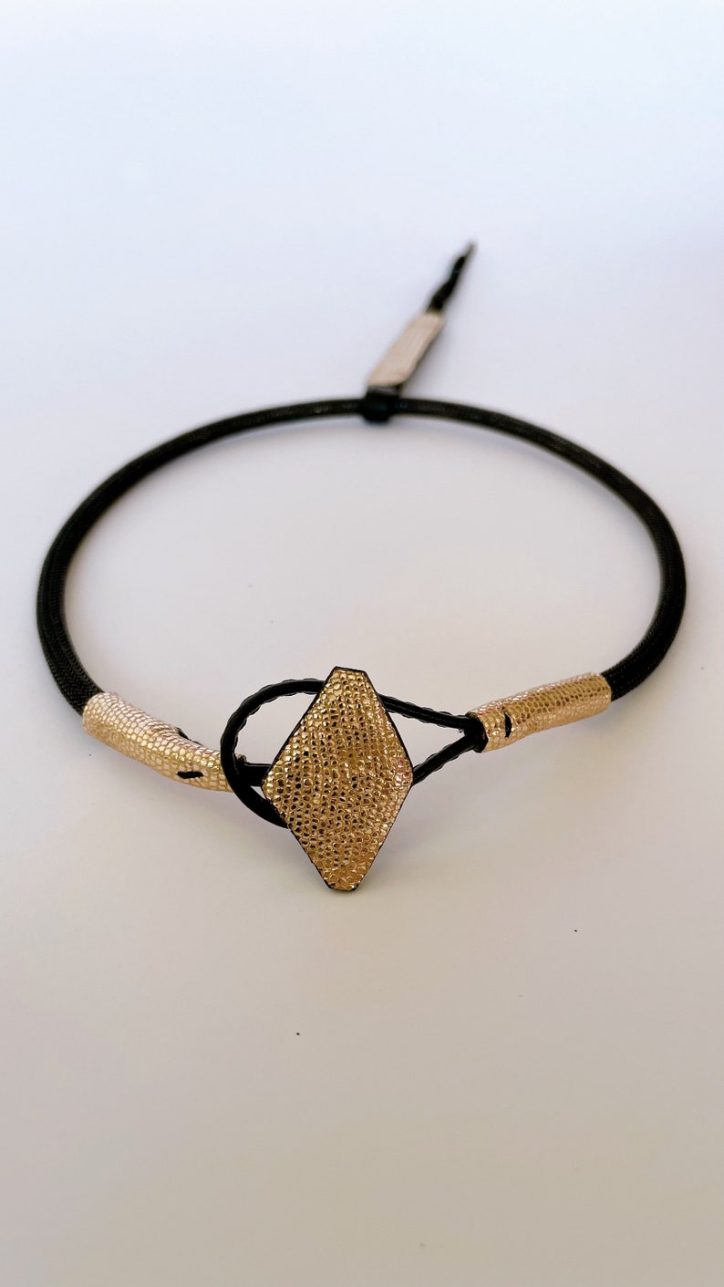 May include: A black cord bracelet with a gold diamond-shaped clasp. The clasp has a textured surface.