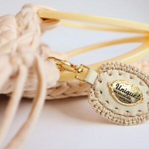May include: A beige crocheted bag with a cream-colored leather tag that says "Unique! by Wioletta Szylypko" with a small rhinestone.