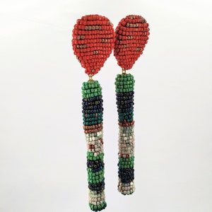 May include: A pair of long, beaded earrings with a red, green, white, and black color scheme. The earrings are made of small, round beads that are strung together to create a long, cylindrical shape. The earrings are finished with a small, red bead at the top.