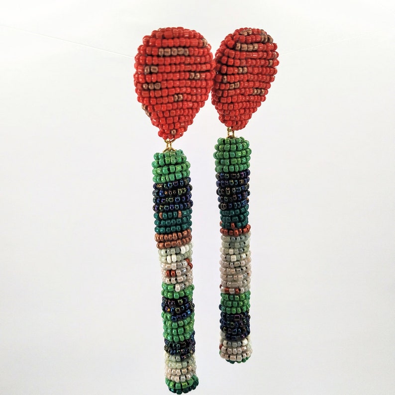 May include: A pair of long, beaded earrings with a red, green, white, and black color scheme. The earrings are made of small, round beads that are strung together to create a long, cylindrical shape. The earrings are finished with a small, red bead at the top.
