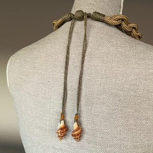May include: A brown and green rope necklace with two shell pendants. The necklace is tied with a knot at the back of the neck.