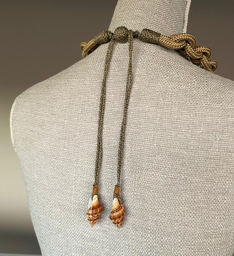 May include: A brown and green rope necklace with two shell pendants. The necklace is tied with a knot at the back of the neck.